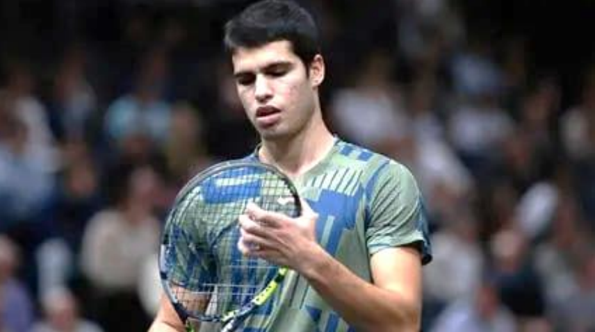 Junger Mann mit Tennisschläger und Ball, schaut auf den Ball, unscharfes Publikum im Hintergrund.
